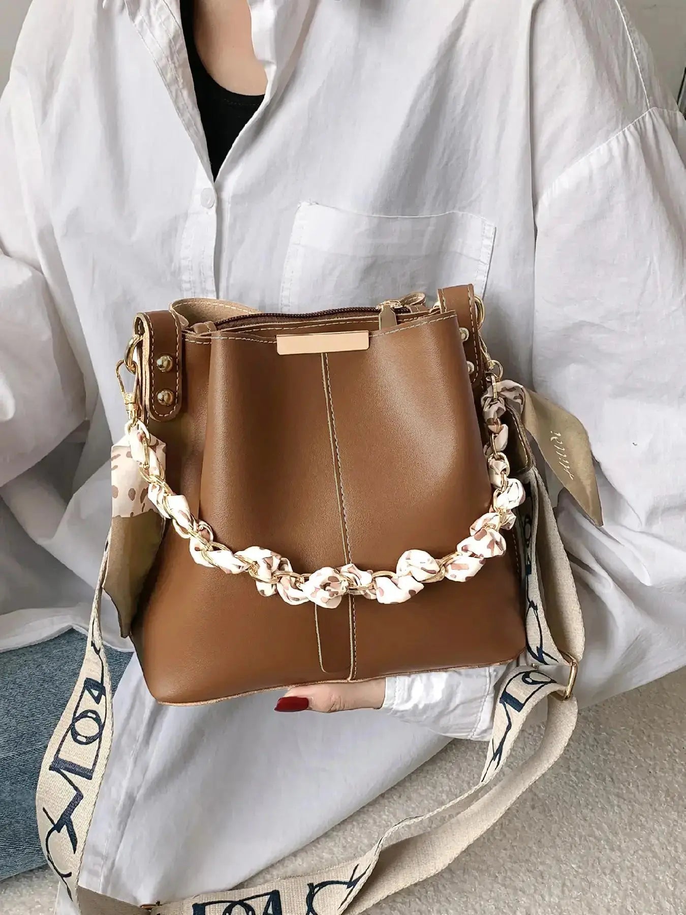 Brown PU leather bucket crossbody bag held by model wearing white oversized shirt and jeans, gold-tone chain and cream leopard print silk scarf handle, cream and black letter-print wide crossbody strap, gold rectangular hardware plate, close-up front view