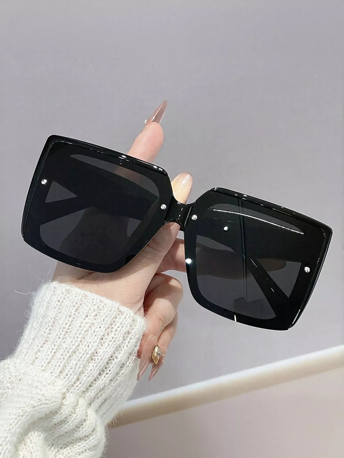 Black oversized square sunglasses with dark lenses and small silver-tone front accents, held by a manicured hand
