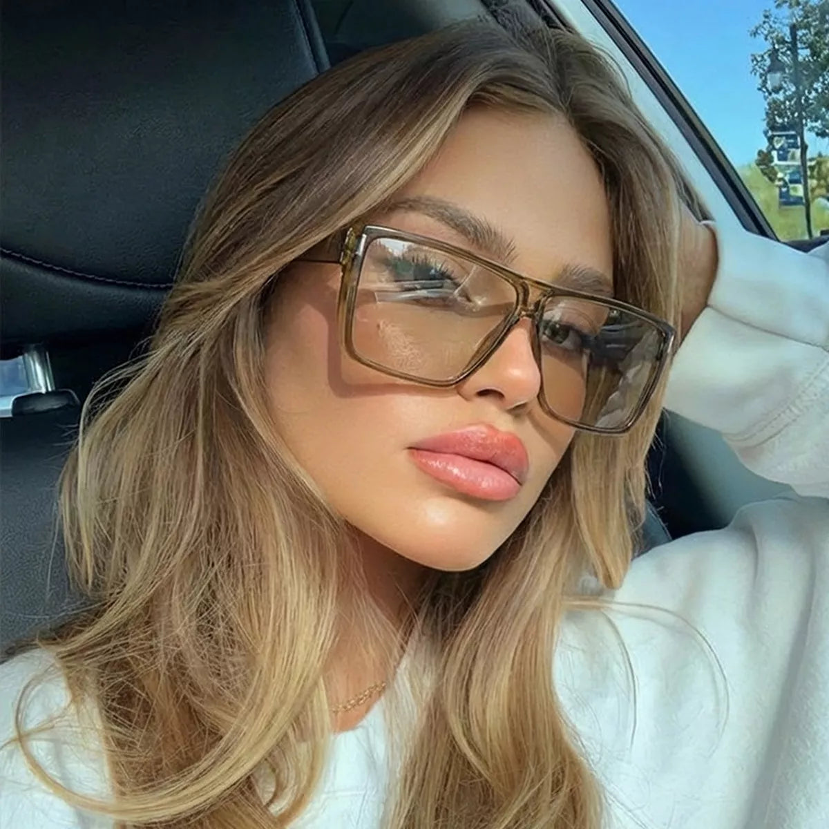 Model seated in a car wearing translucent brown oversized square sunglasses and a white sweatshirt