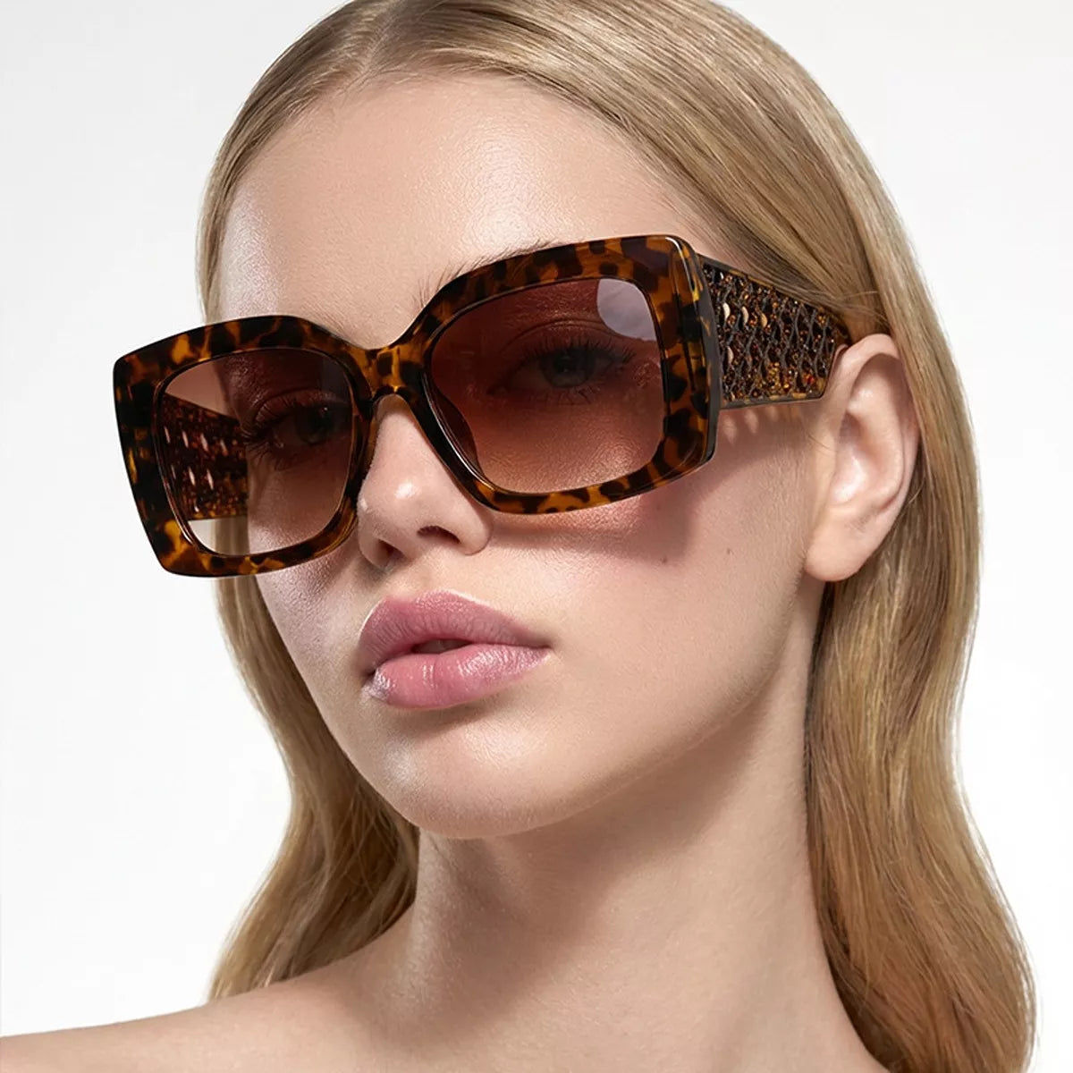 Blonde model wearing amber-brown tortoiseshell oversized square sunglasses with brown gradient lenses and openwork lattice temples against a white background