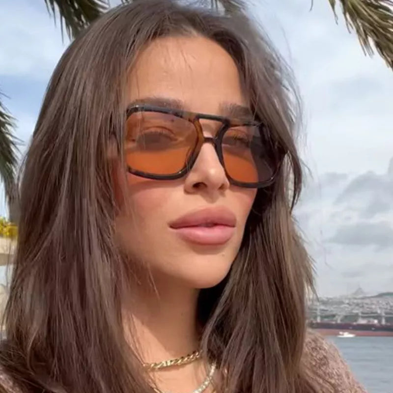 Woman wearing oversized tortoiseshell square sunglasses with warm brown lenses and layered necklaces, with palm fronds and water in the background