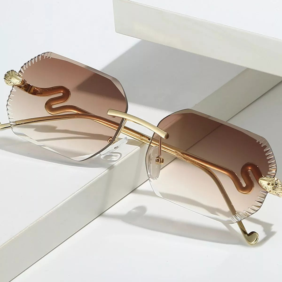 Brown gradient rimless sunglasses with clipped-corner lenses, gold-tone snake temples, and clear nose pads, resting on white display blocks