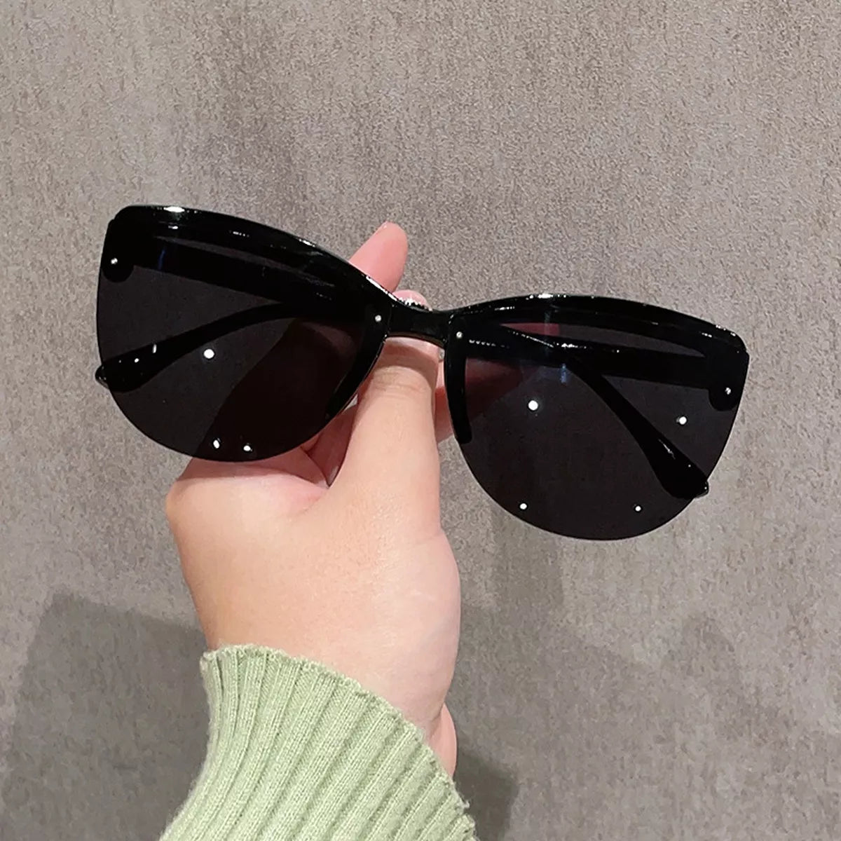 Black oversized semi-rimless cat-eye sunglasses with dark lenses, held by a hand in a pale green ribbed sleeve against a textured gray background