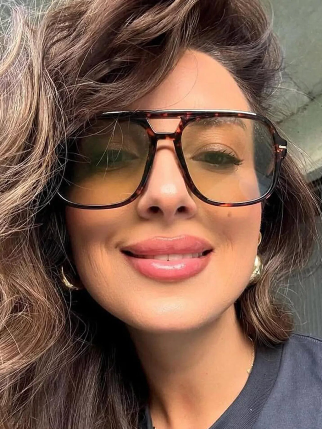 Smiling model wearing oversized brown tortoiseshell-look square aviator sunglasses with warm brown-tinted lenses and a double bridge, gold-tone earrings and a dark top