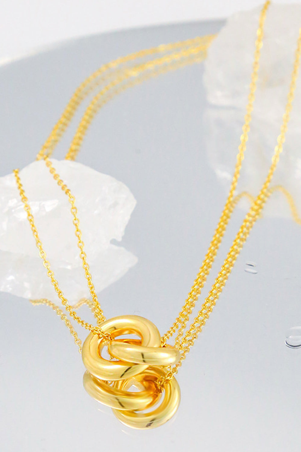 Gold-tone interlocking ring pendant necklace on a mirrored surface beside a white decorative stone, with the pendant reflected below