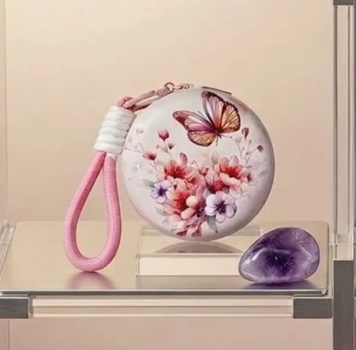 Round hard-shell compact mirror keychain with white case featuring an orange and pink butterfly above pink and lavender floral print, attached to a pink rope wrist strap with white wrapped section and gold carabiner clip, displayed on a clear acrylic stand beside an amethyst crystal on a warm beige surface.