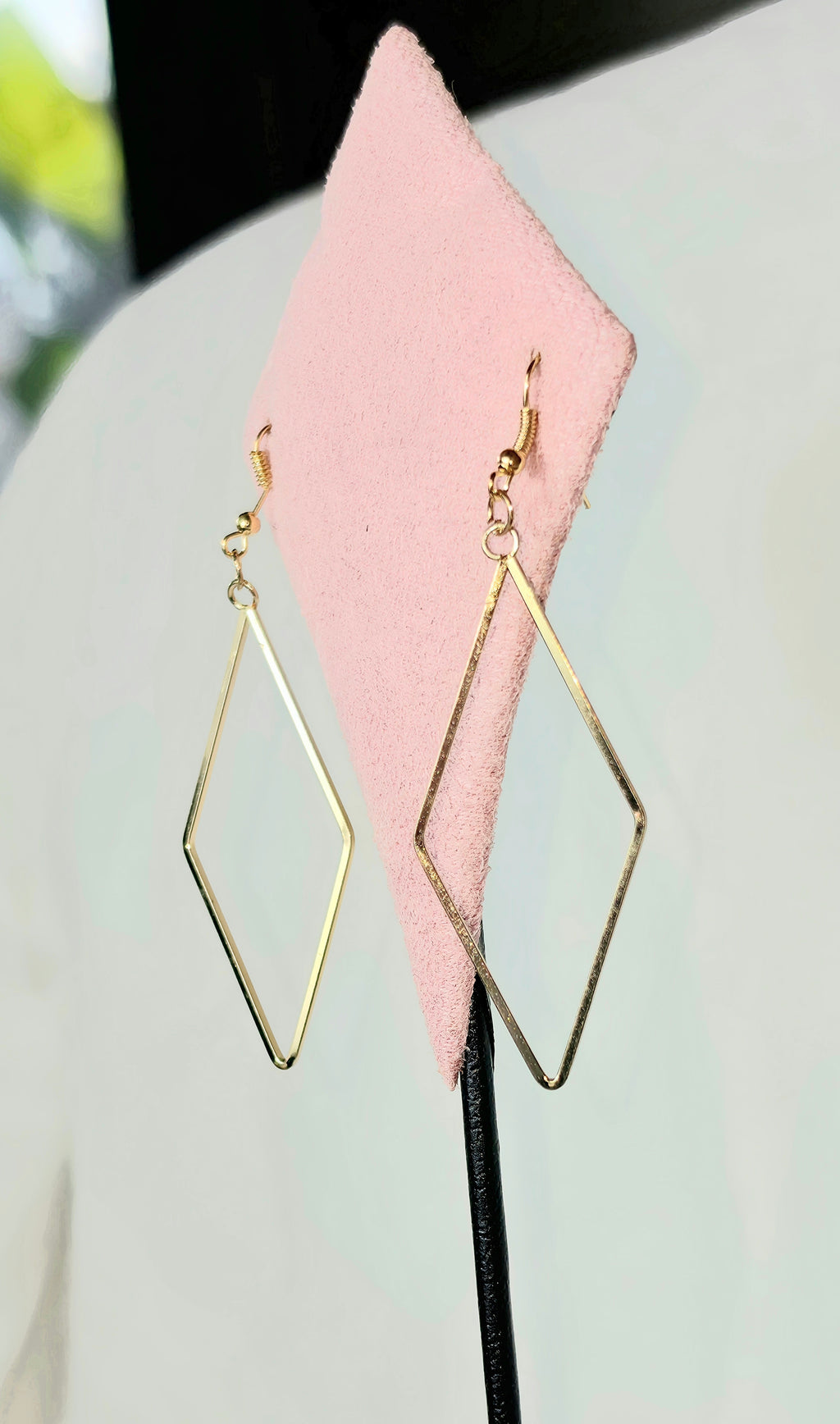 Pair of gold open diamond drop earrings hanging from a pink diamond-shaped suede jewelry display stand, showing slim gold wire rhombus shape and fish hook ear wire, soft gray background