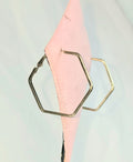 Hexagonal hoop earrings in silver and gold-tone metals, shown hanging against a pale light blue background with a pink fabric prop.