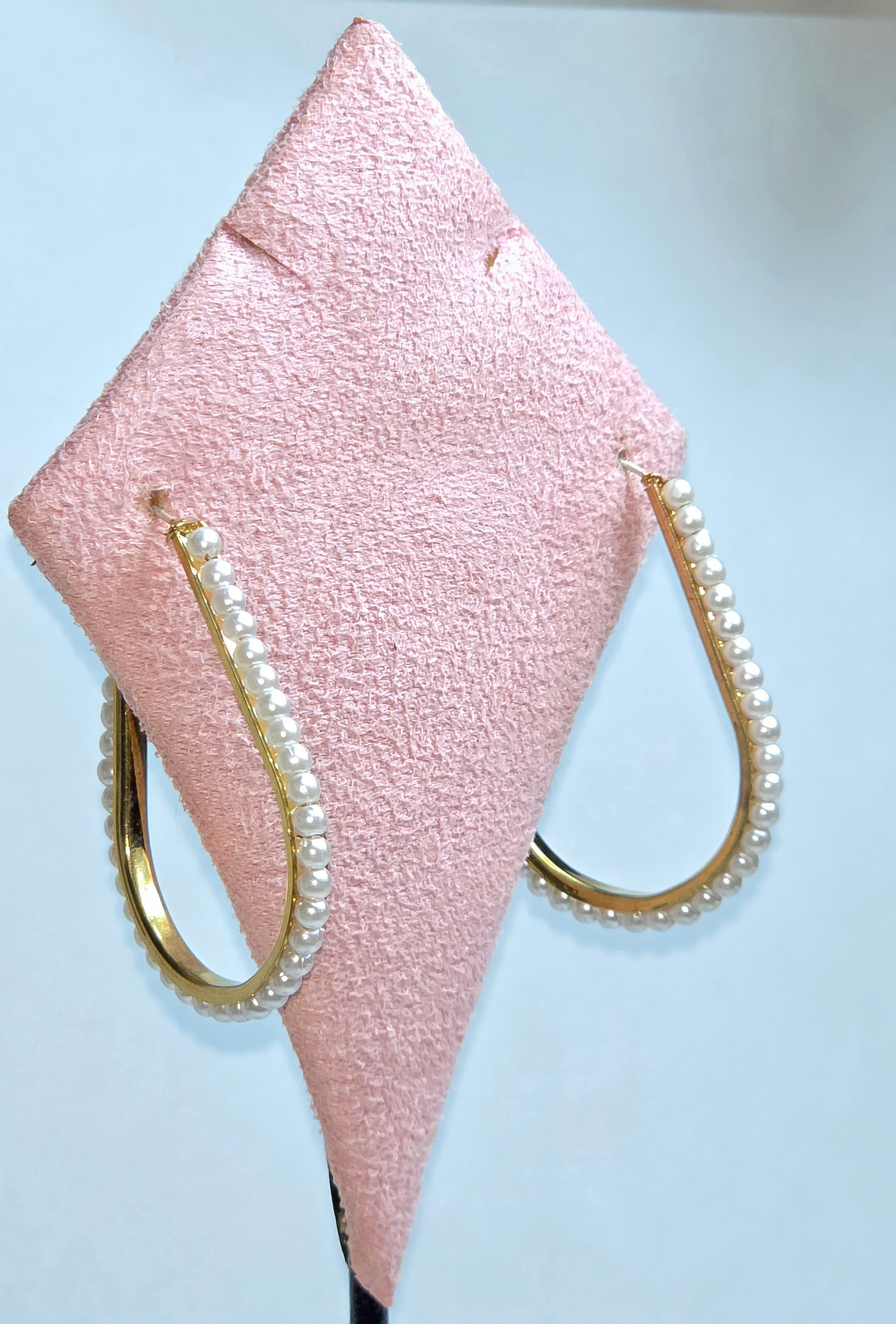 A pair of gold hoop earrings decorated with white faux pearls, displayed on a pink textured jewelry stand against a light blue background.