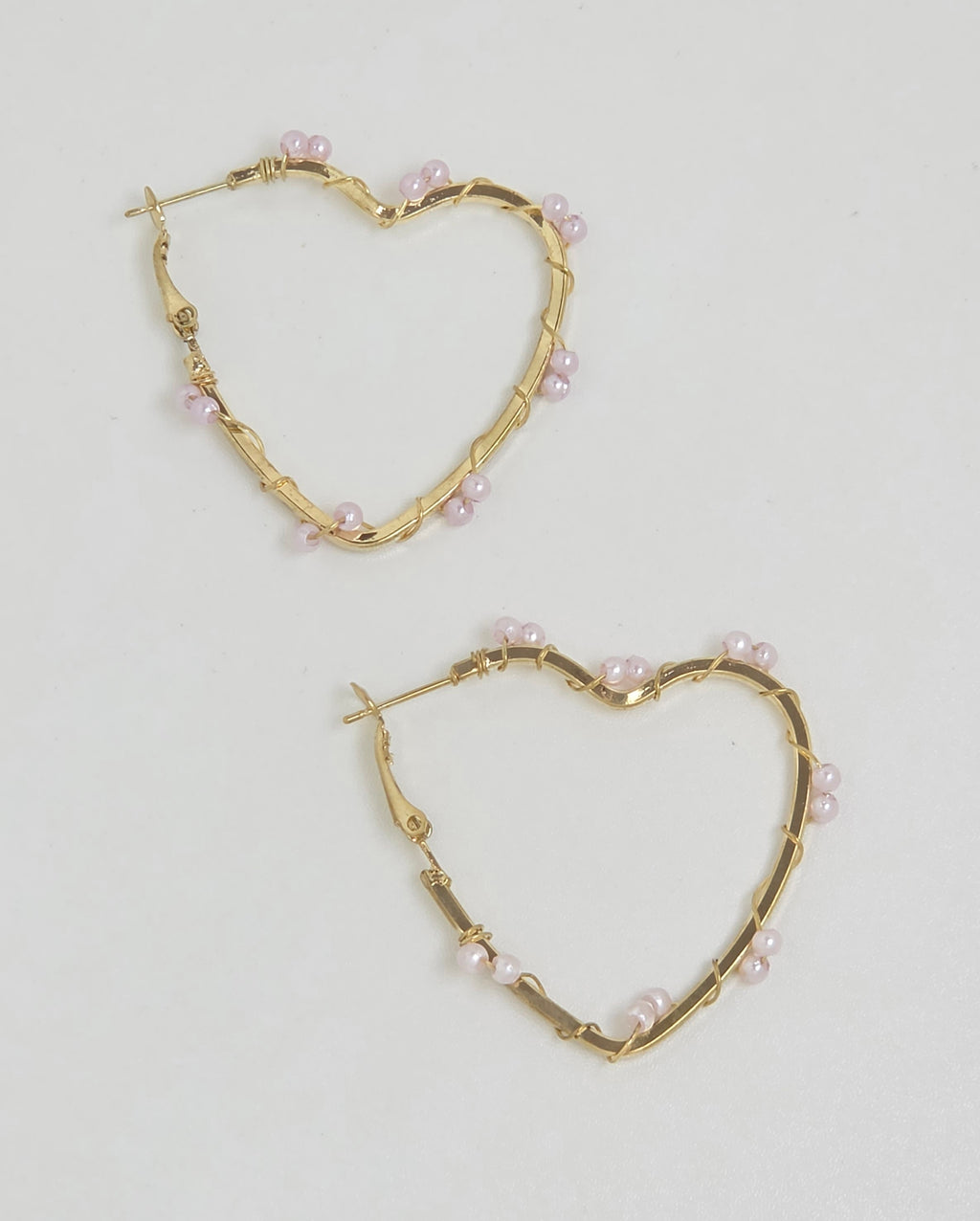 A pair of gold heart-shaped hoop earrings decorated with small pink pearls, displayed on a clean white background.