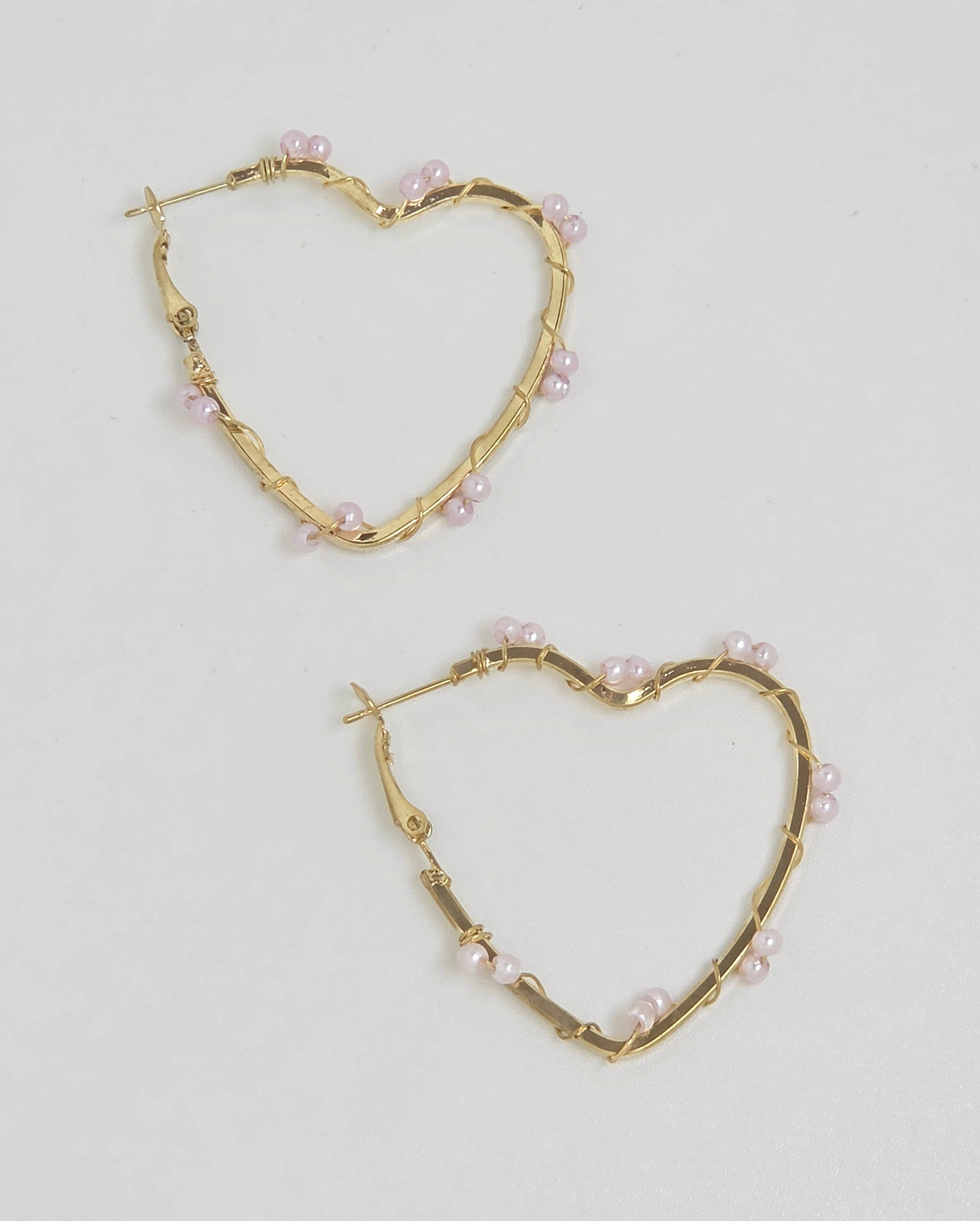 A pair of gold heart-shaped hoop earrings decorated with small pink pearls, displayed on a clean white background.