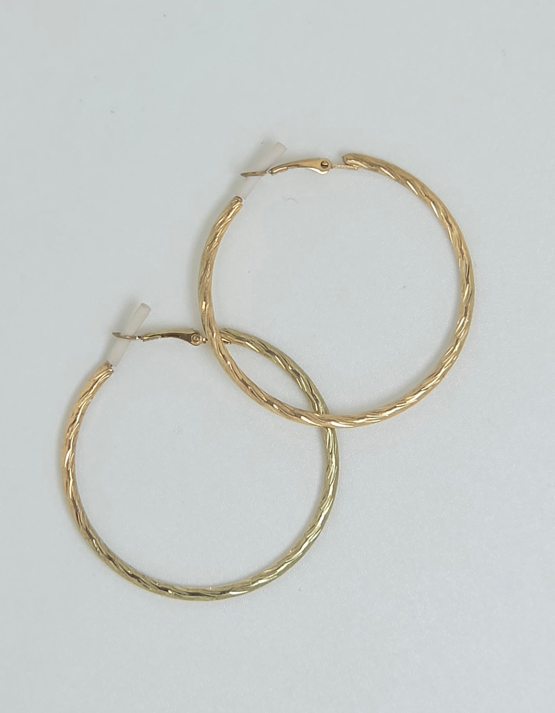 A pair of gold hoop earrings with a textured, twisted finish, displayed on a plain light gray background.