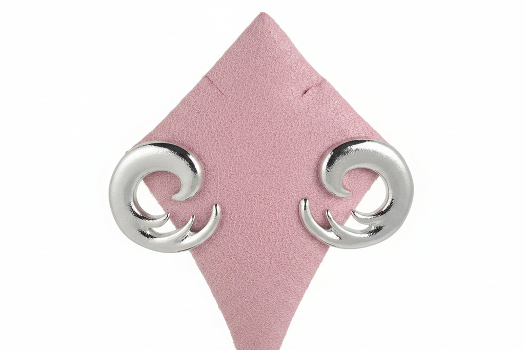Pair of bold silver spiral swirl stud earrings displayed on pink diamond-shaped suede jewelry stand, showing chunky open spiral design in high-polish silver-tone metal, gray background