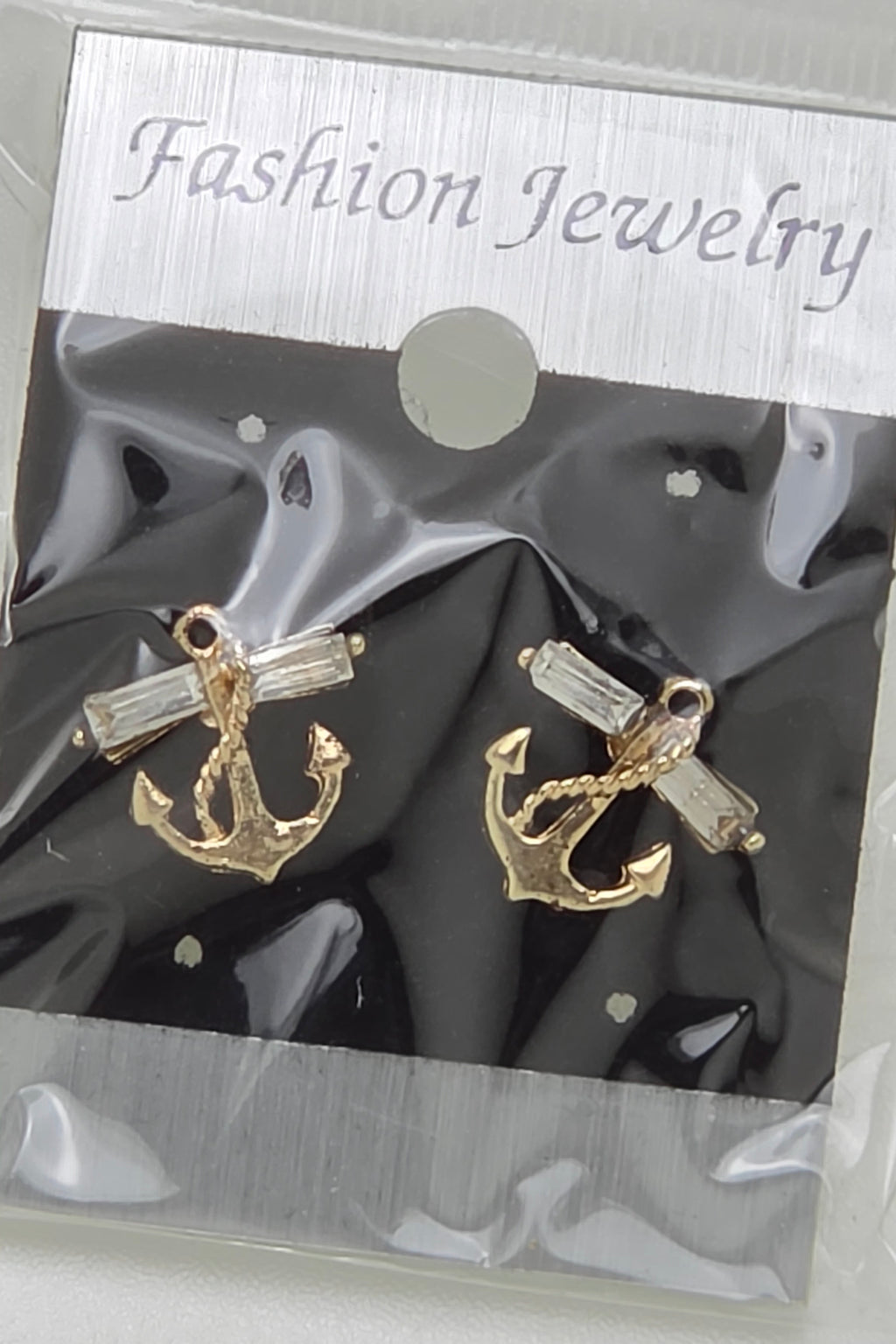 A pair of gold anchor stud earrings with clear rectangular crystals, presented on a black backing card inside a clear plastic package with a silver header that reads 'Fashion Jewelry'.