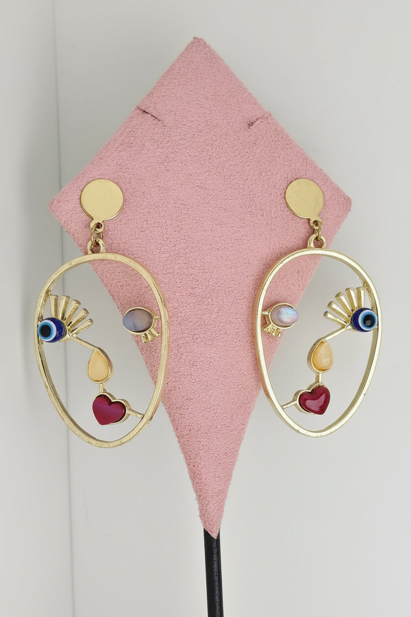 A pair of gold-toned dangle earrings displayed on a pink jewelry stand against a white background. Each earring has an oval frame with a teardrop pendant, a small fan motif, and colorful enamel details including blue, purple, and red.