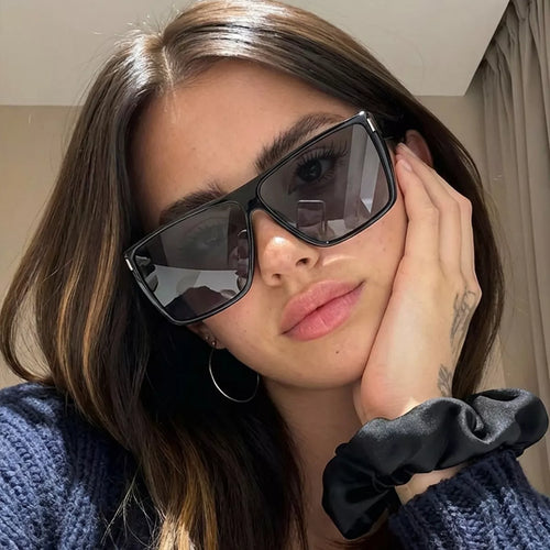 Model wearing black oversized square sunglasses with dark lenses, a dark knit top, and a black wrist scrunchie