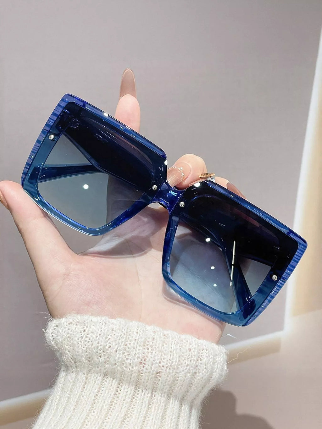 Front view of blue oversized square sunglasses with gradient lenses, held by a manicured hand in a white knit sleeve