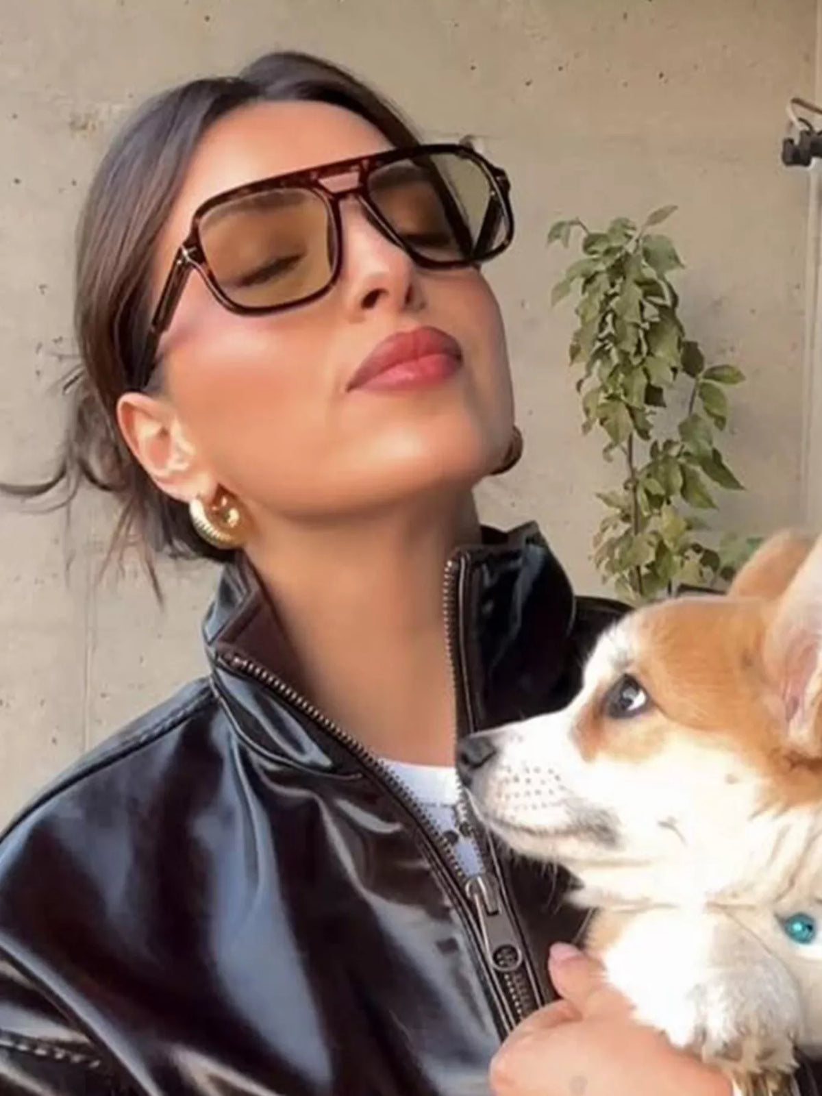 Model wearing oversized brown tortoiseshell-look aviator sunglasses with warm tinted lenses and a double bridge, gold-tone earrings and a glossy black jacket while holding a tan-and-white dog