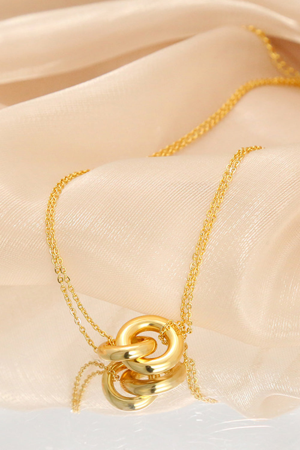 Gold-tone linked-ring pendant necklace draped over peach fabric and a reflective surface, with the pendant reflected below