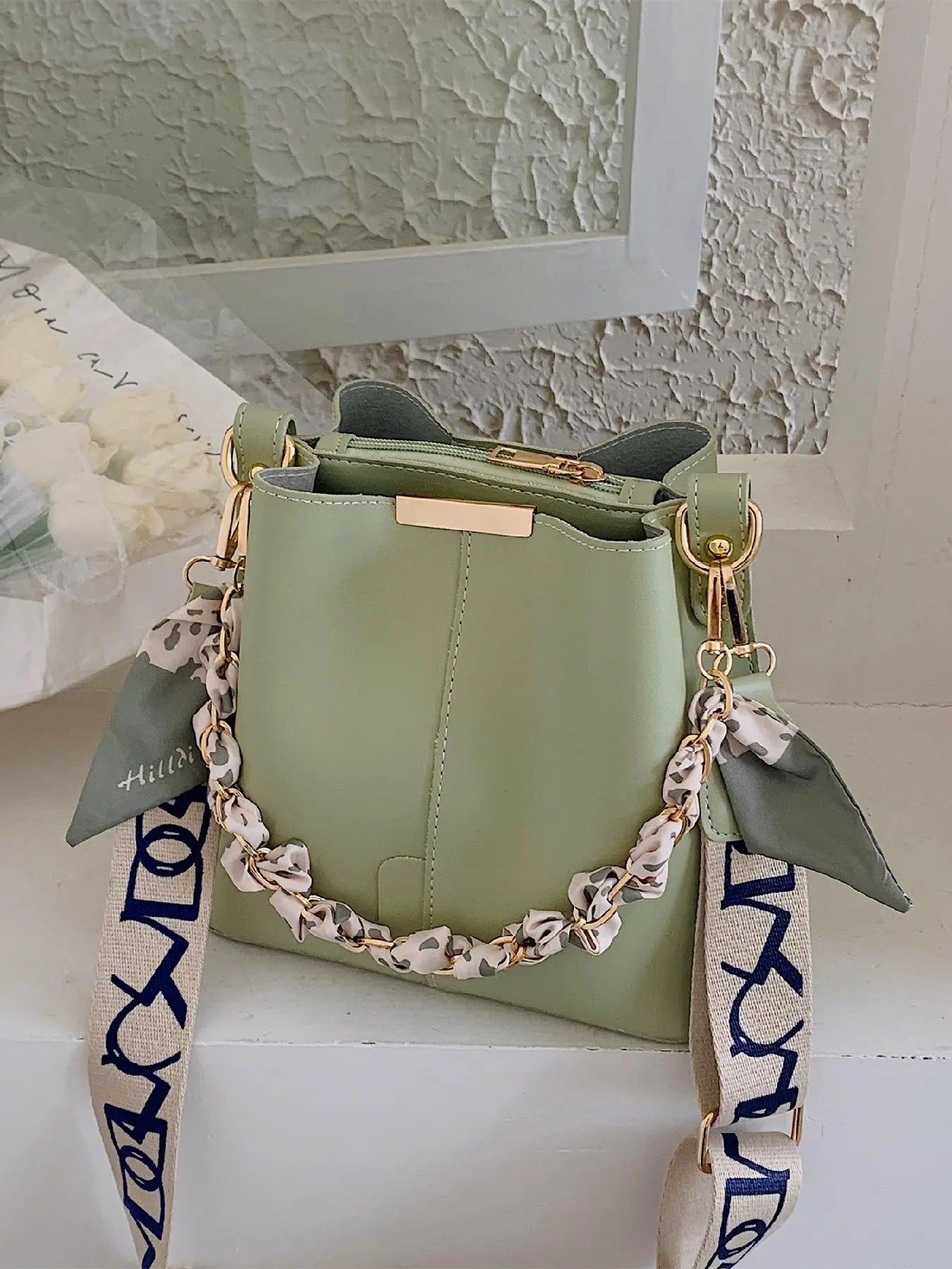 Sage green PU leather bucket crossbody bag with gold-tone chain and cream leopard print silk scarf handle, cream and navy blue letter-print wide crossbody strap, gold rectangular hardware plate, styled on white shelf with textured plaster wall and white flowers in background