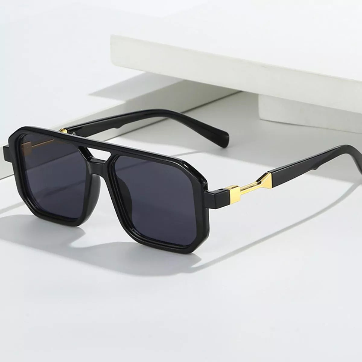 Black oversized square sunglasses with dark lenses, a double bridge and gold-tone temple accents displayed with open arms on a white surface