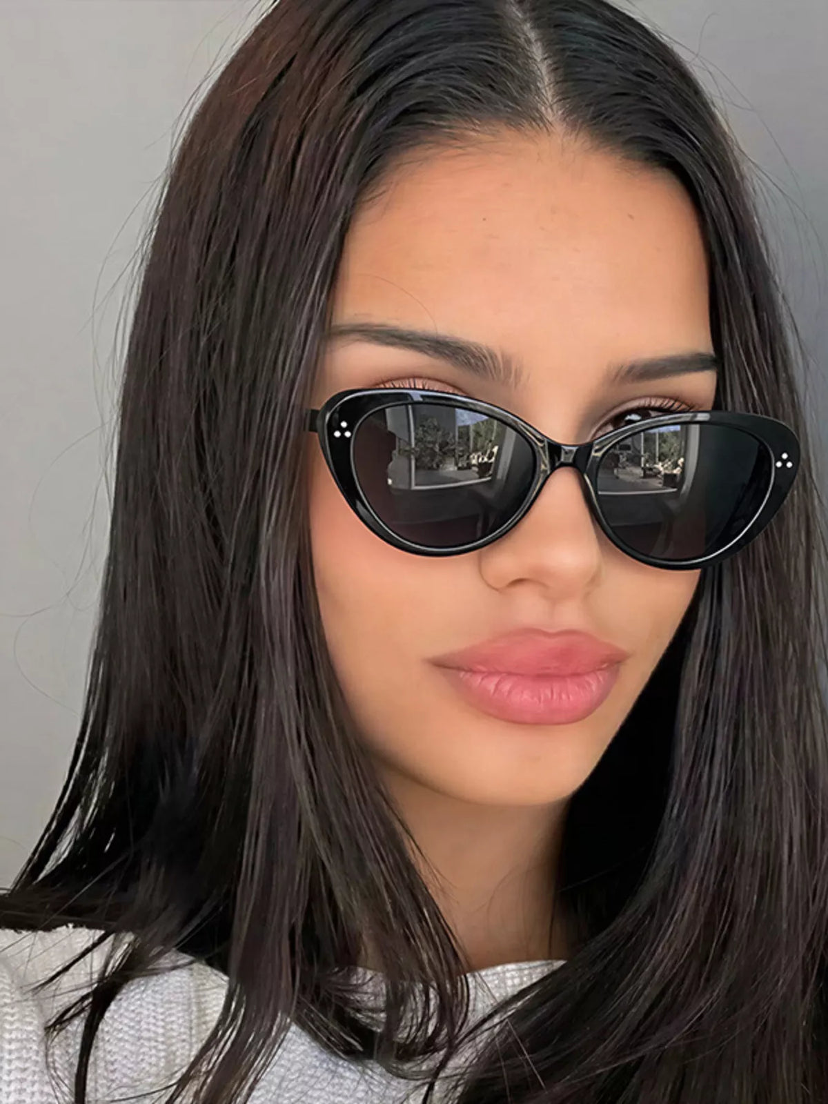 Model wearing glossy black cat-eye sunglasses with dark lenses and three-dot corner accents, styled with a light-colored top