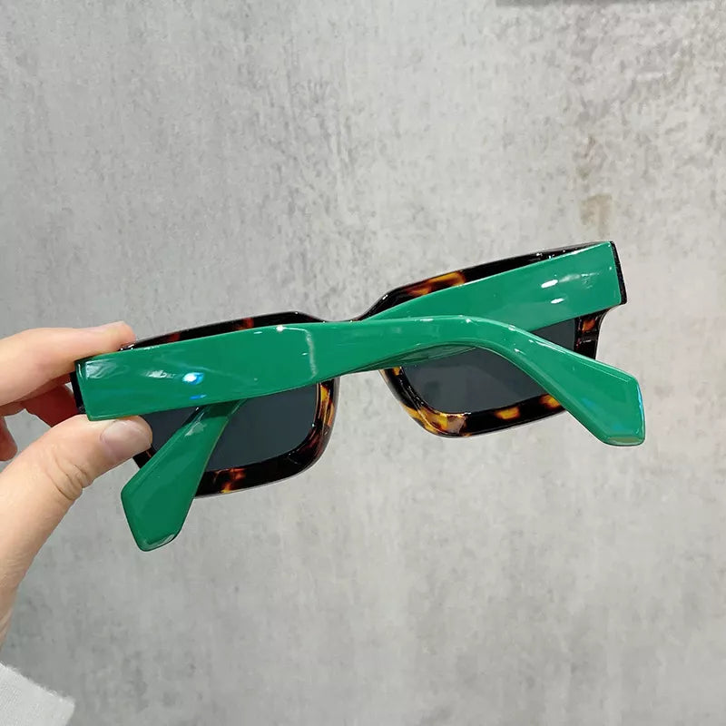 Rear view of chunky rectangular sunglasses with glossy green temples, a tortoiseshell front, and dark lenses, held against a gray textured wall