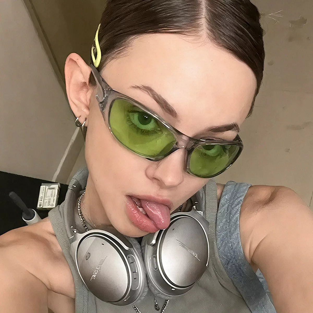 Side-angle portrait of a model wearing green-tinted rectangular sunglasses with a translucent smoky-gray frame and headphones around her neck