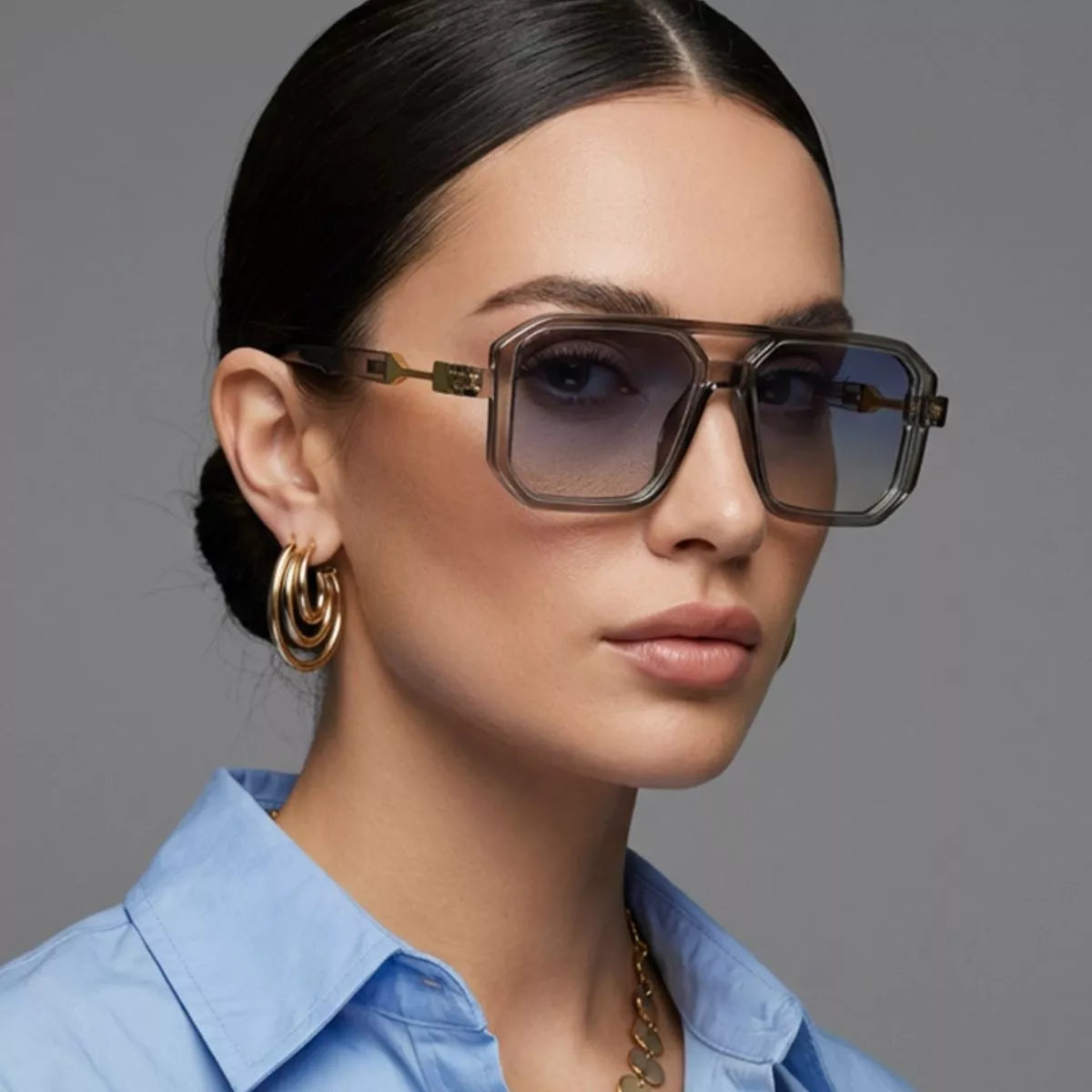 Model wearing smoky-gray oversized square sunglasses with gradient lenses, gold-tone temple details, gold hoop earrings and a blue collared shirt against a gray background