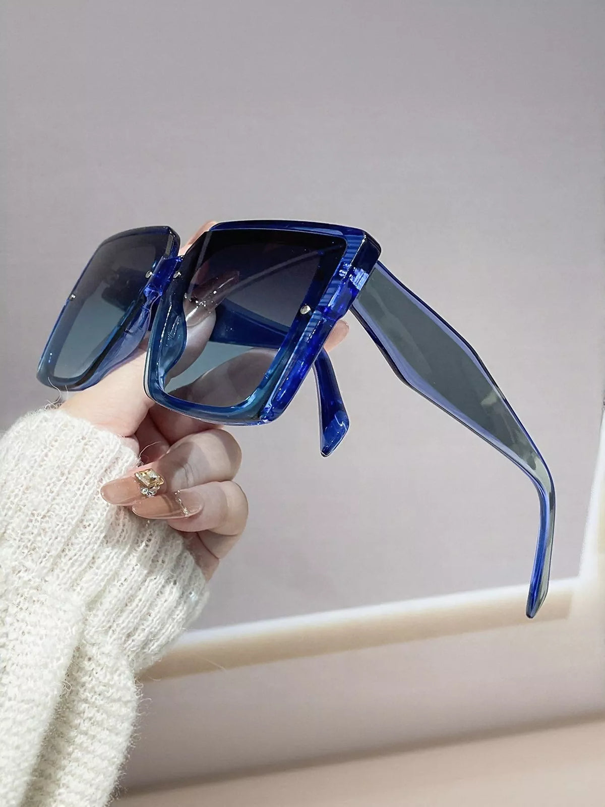 Side view of blue square sunglasses showing wide temples and gradient lenses, held by a hand in a white knit sleeve
