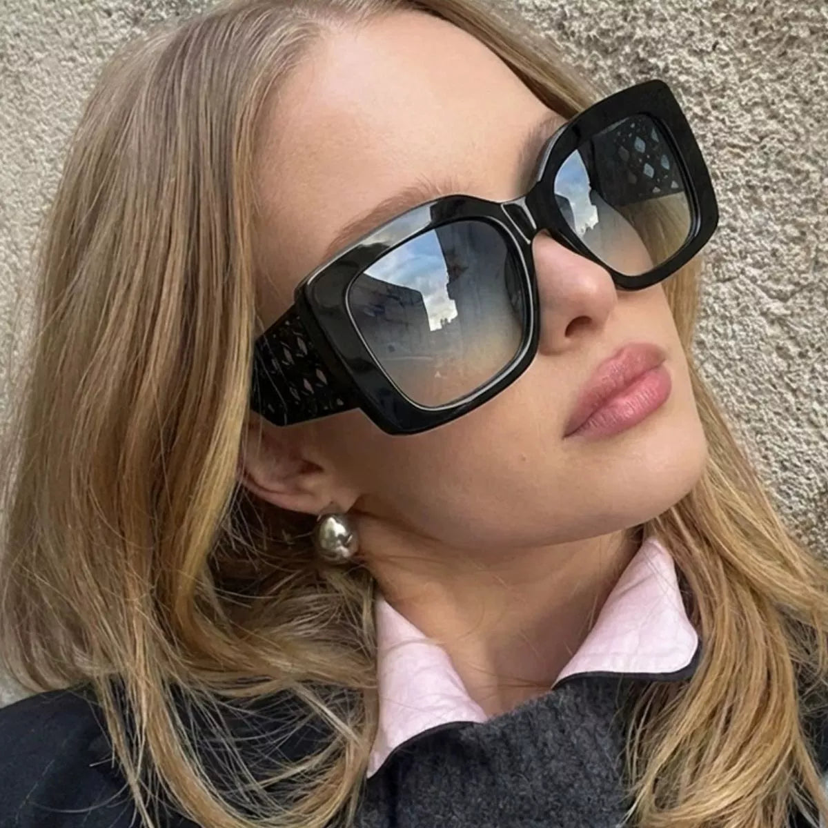 Blonde model wearing black oversized square sunglasses with gray gradient lenses and lattice temples, a dark sweater over a pink collared shirt and a silver-tone earring beside a textured wall