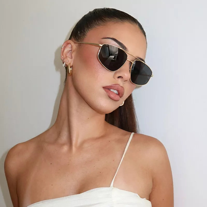 Model wearing gold-tone metal aviator sunglasses with dark lenses and a double bridge, styled with gold-tone hoop earrings and a white top