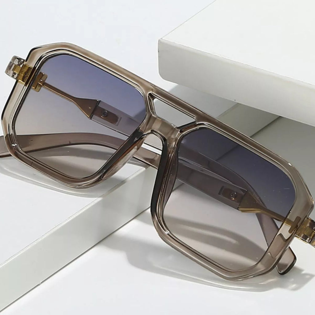 Oversized translucent smoky-gray square sunglasses with gradient lenses, a double bridge and gold-tone temple details resting on white display blocks