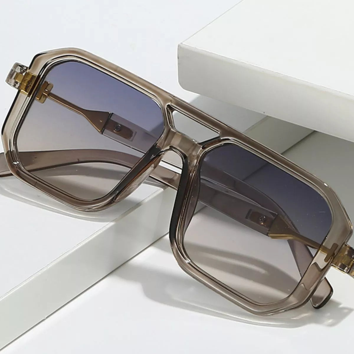 Oversized translucent smoky-gray square sunglasses with gradient lenses, a double bridge and gold-tone temple details resting on white display blocks