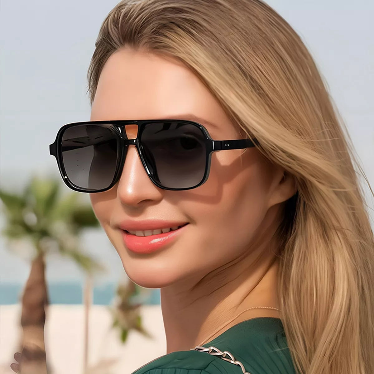 Blonde model wearing black oversized aviator sunglasses with dark lenses and a cutout bridge, styled with a green top and chain shoulder strap near palm trees