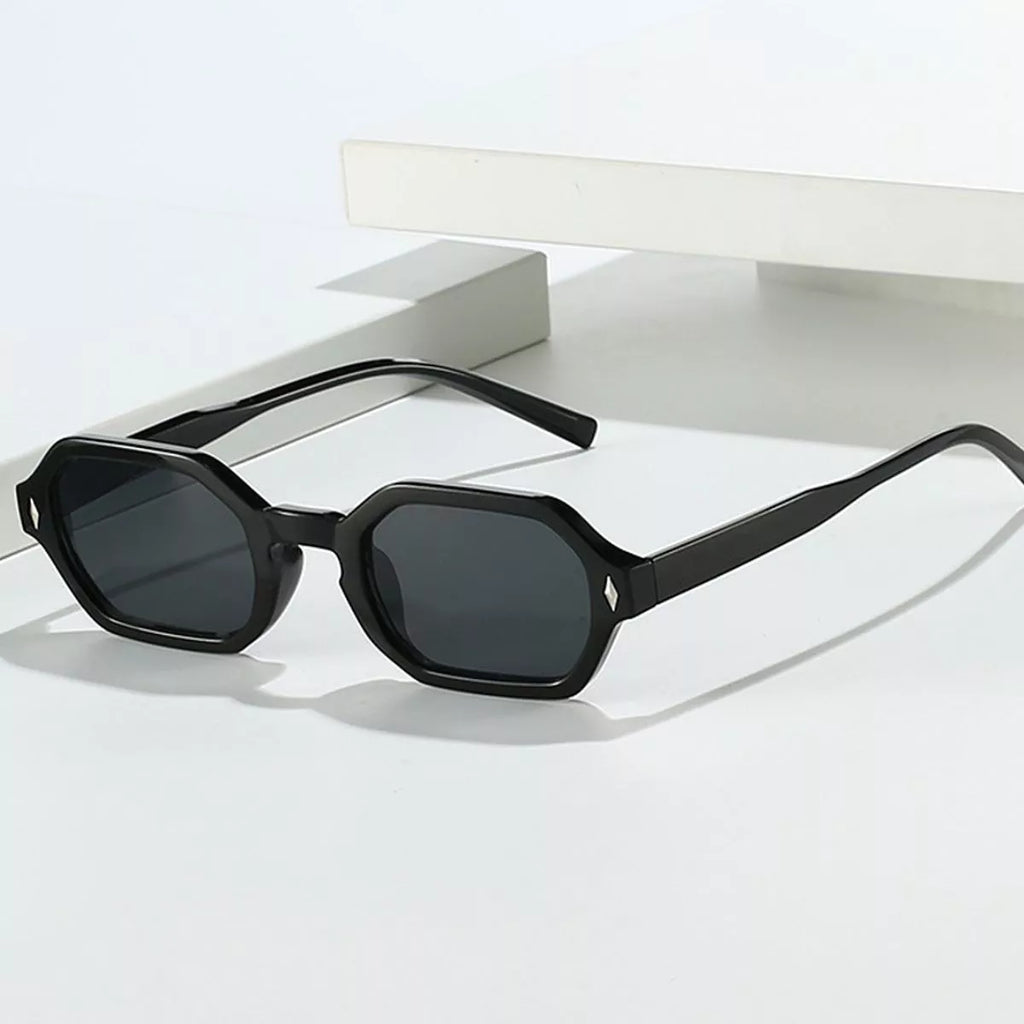 Black geometric sunglasses with dark gray lenses and silver-tone diamond-shaped corner accents, held against a white card by a hand in a metallic silver sleeve