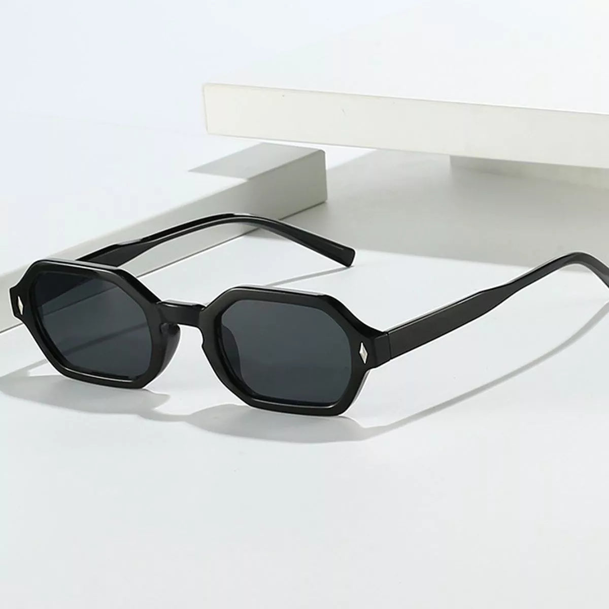 Black geometric sunglasses with dark gray lenses and silver-tone diamond-shaped corner accents, held against a white card by a hand in a metallic silver sleeve