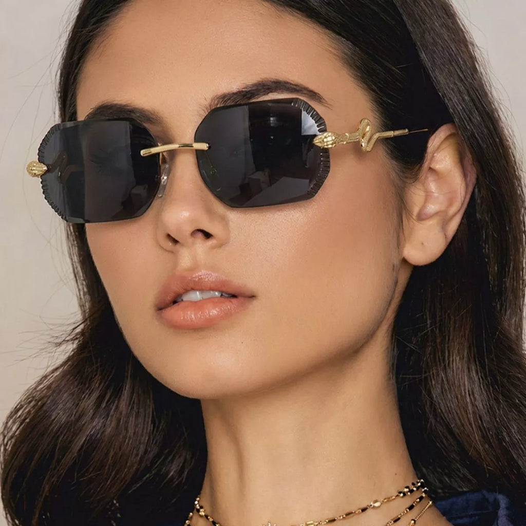 Model wearing dark rimless geometric sunglasses with gold-tone snake temples, styled with layered delicate necklaces