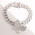 Overhead angled flat lay of a fully pave clear rhinestone Miami Cuban chain link bracelet in rhodium silver tone on a white angled surface. An oversized fully pave clear rhinestone butterfly charm measuring 1.25 inches is centered at the bottom of the bracelet. A rhinestone-crusted square push-lock box clasp with a hinged safety bar is visible at the top.