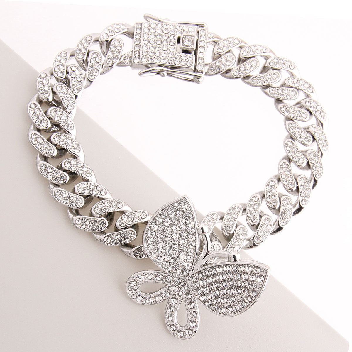 Overhead angled flat lay of a fully pave clear rhinestone Miami Cuban chain link bracelet in rhodium silver tone on a white angled surface. An oversized fully pave clear rhinestone butterfly charm measuring 1.25 inches is centered at the bottom of the bracelet. A rhinestone-crusted square push-lock box clasp with a hinged safety bar is visible at the top.