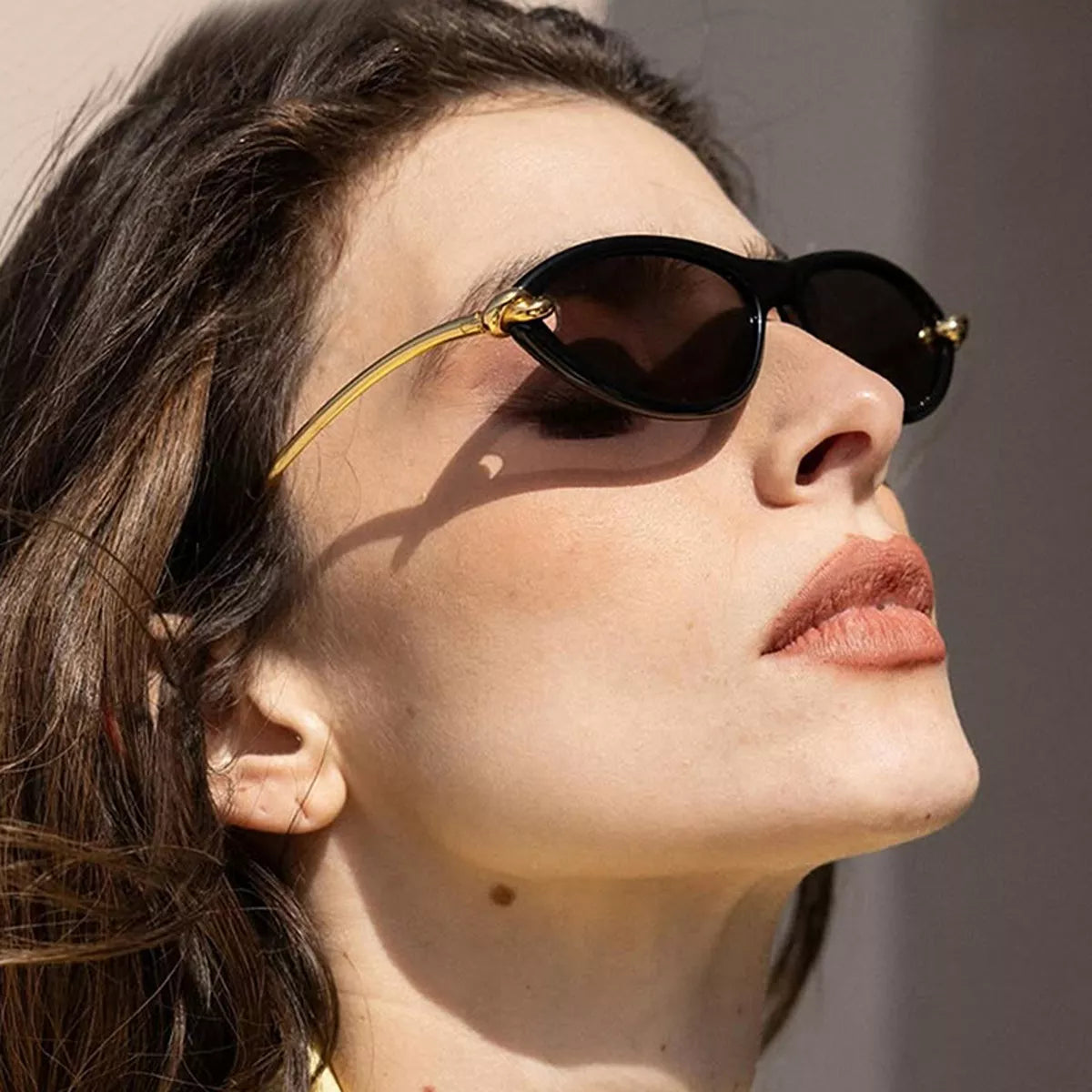 Side-profile close-up of a model wearing black cat-eye sunglasses with dark lenses and gold-tone knot details in sunlight