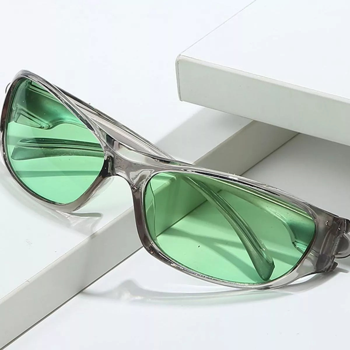 Green lens rectangular sunglasses with a translucent smoky-gray polycarbonate frame displayed on white geometric blocks
