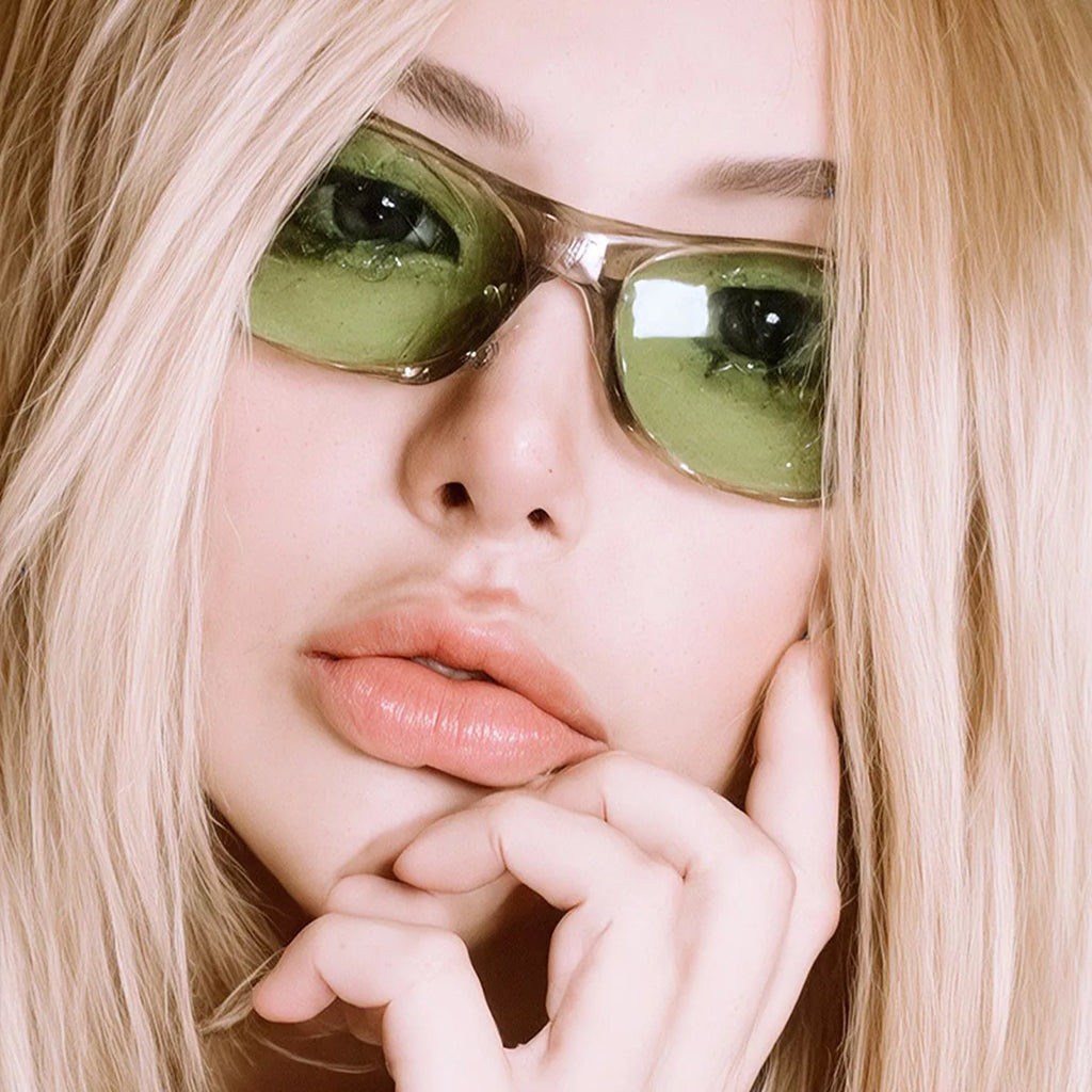 Close-up of a blonde model wearing green-tinted rectangular sunglasses with a translucent smoky-gray frame, resting her hand against her chin