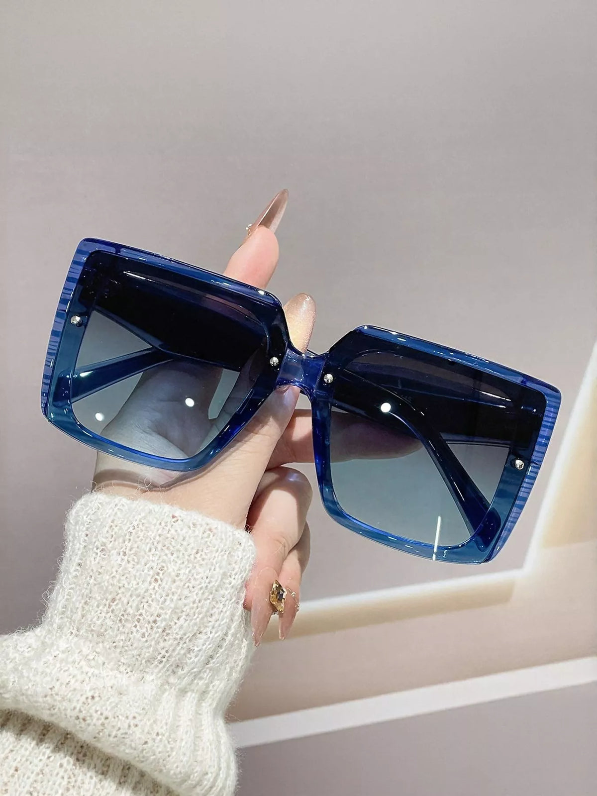 Blue translucent oversized square sunglasses with blue-gray gradient lenses, resting across an open hand against a gray wall