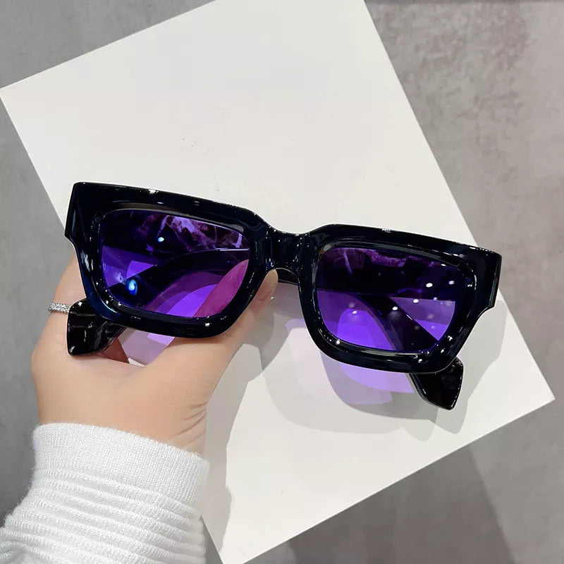 Glossy black rectangular sunglasses with chunky frames and purple lenses, held over a white sheet against a gray background