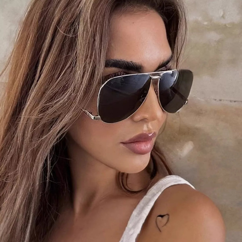 Close-up of a woman in silver-tone aviator sunglasses with dark teardrop lenses, long brown hair, and a white top against a beige wall