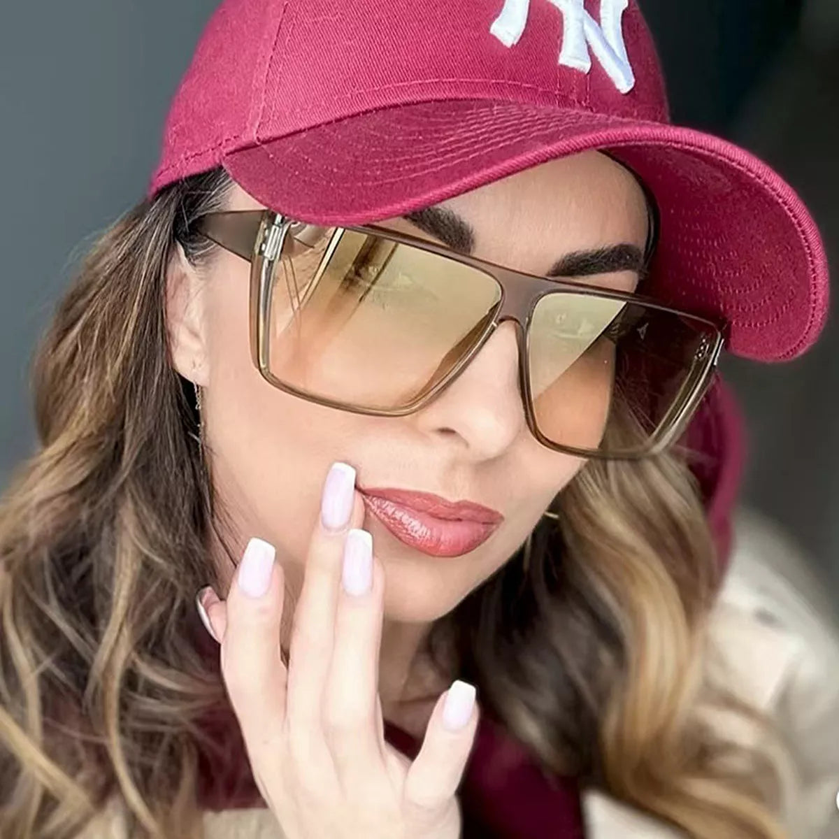 Model wearing translucent brown oversized square sunglasses with warm tinted lenses and a burgundy baseball cap