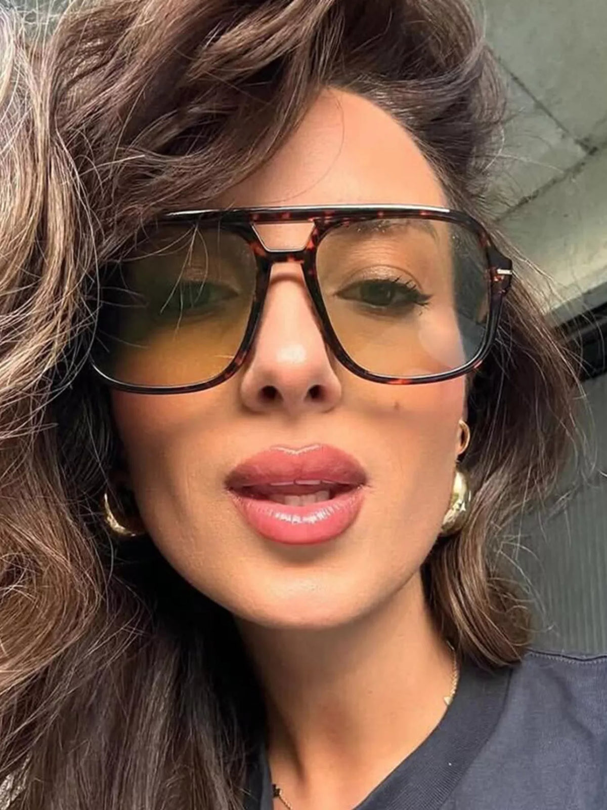Close-up of a model with parted lips wearing oversized brown tortoiseshell-look aviator sunglasses with warm tinted lenses, a double bridge and gold-tone earrings