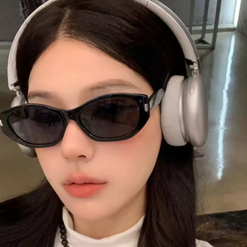 Front close-up of a woman wearing black cat-eye sunglasses with dark lenses and silver-tone corner accents, paired with silver over-ear headphones and a white top