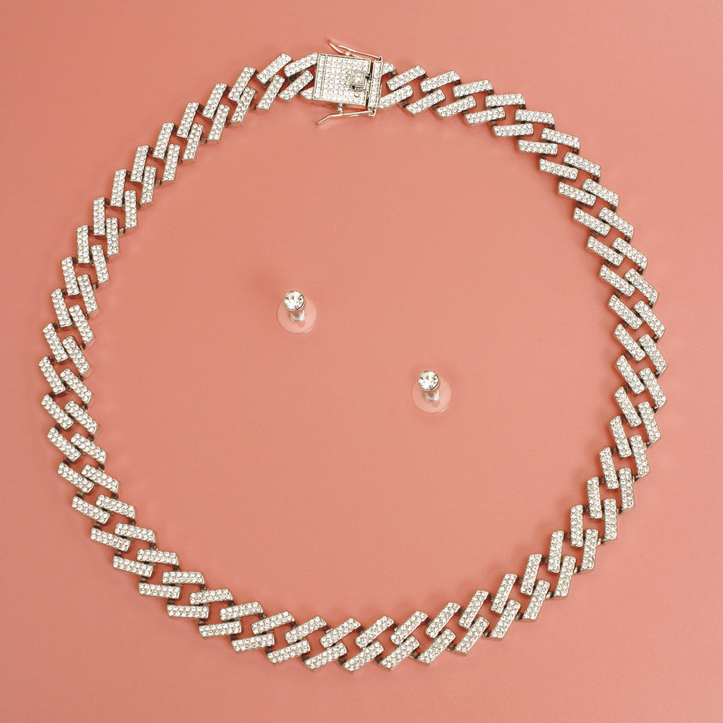 Overhead flat lay of a wide silver tone fully pave clear rhinestone square Cuban chain link necklace arranged in a circle on a coral pink surface. A square push-lock box clasp covered in clear rhinestones is visible at the top. Two small clear rhinestone round stud earrings with clear rubber butterfly post backings are displayed inside the necklace loop.